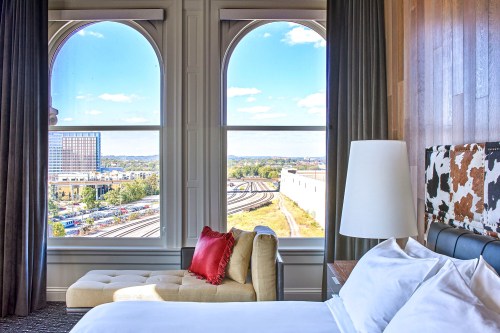 A hotel room with a bed and a view of train tracks and buildings through large arched windows.