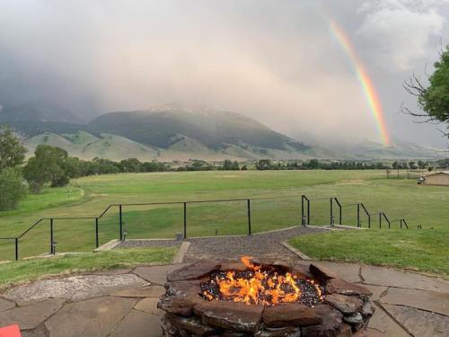 A scenic view of a rainbow over mountains, with a fire pit in the foreground and a grassy field below.