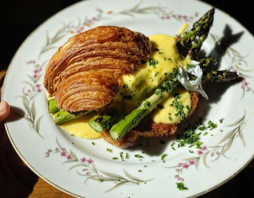 A croissant topped with asparagus and drizzled with hollandaise sauce, garnished with herbs on a decorative plate.