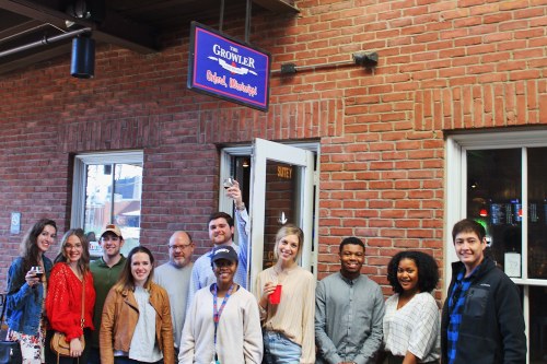A group of diverse people poses together outside a restaurant named "The Growler," smiling and holding drinks.