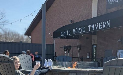 A woman relaxes in an outdoor seating area at White Horse Tavern, holding a drink near a fire pit.