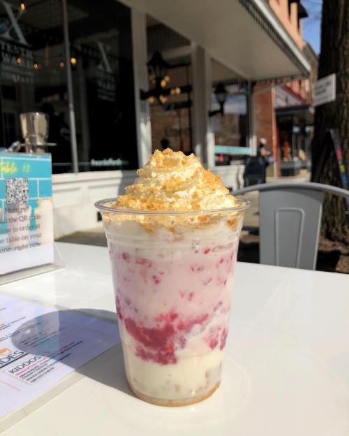 A clear cup filled with layers of creamy dessert, topped with whipped cream and crumbled cookies, set outdoors.