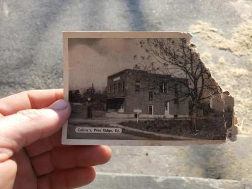 A hand holds a vintage photo of Collier's building in Pine Ridge, Kentucky, with a partially torn corner.