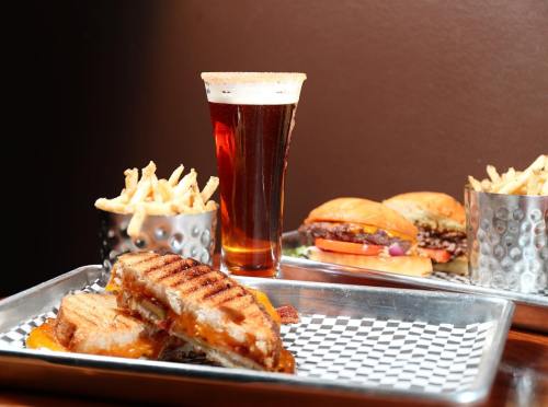 A grilled cheese sandwich, a pint of beer, and sliders with fries on a tray, set against a dark background.