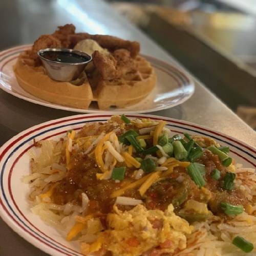 A plate of hash browns topped with cheese, green onions, and salsa, alongside a waffle with fried chicken and syrup.