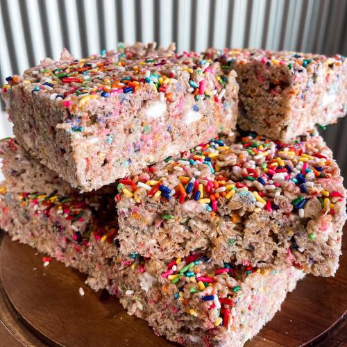 A stack of colorful rice crispy treats topped with rainbow sprinkles on a wooden plate.