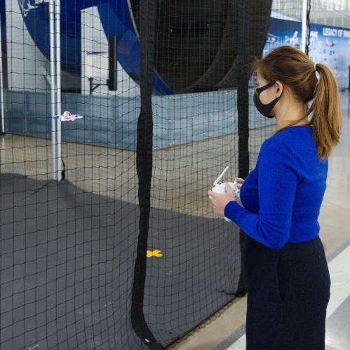 A woman in a blue sweater and mask controls a drone inside a netted area.