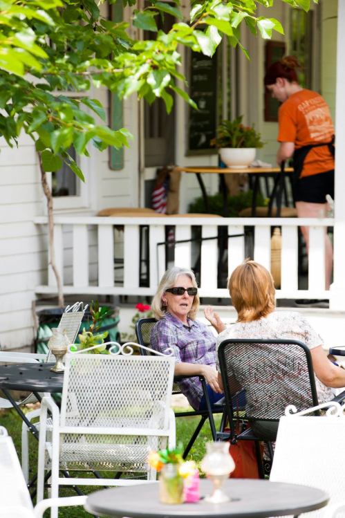 Two women chat at an outdoor café, while a staff member prepares food in the background. Greenery surrounds the scene.
