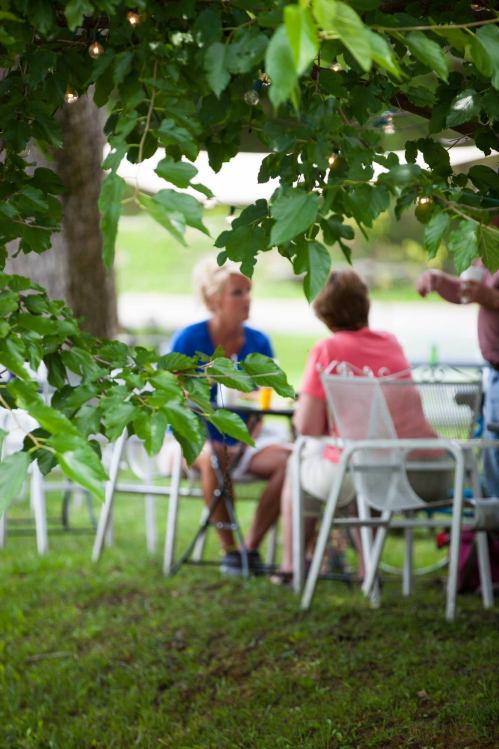 A blurred view of two people sitting at a table under leafy trees, enjoying a casual outdoor gathering.
