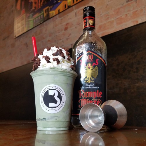 A mint-flavored milkshake topped with whipped cream and chocolate, next to a bottle of Rumple Minze and two jiggers.