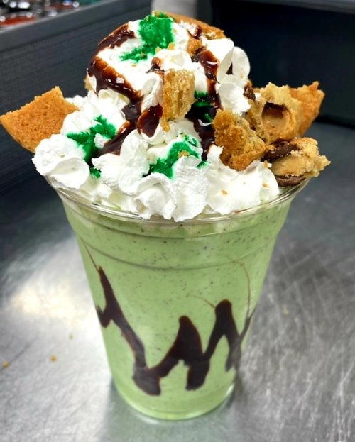A tall cup filled with green mint shake, topped with whipped cream, chocolate drizzle, cookie pieces, and green sprinkles.