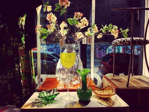 A display of colorful glassware and a vase with flowers, set against a window with a view of parked cars.