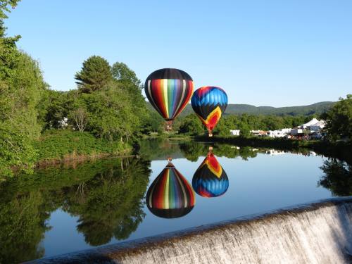 Two colorful hot air balloons float above a calm river, reflecting in the water, surrounded by lush greenery.