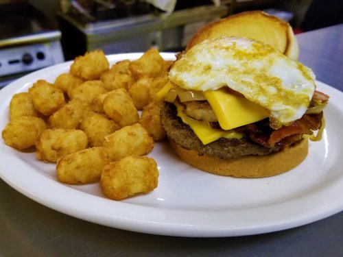 A hearty burger topped with cheese, bacon, and a fried egg, served with a side of crispy tater tots on a white plate.