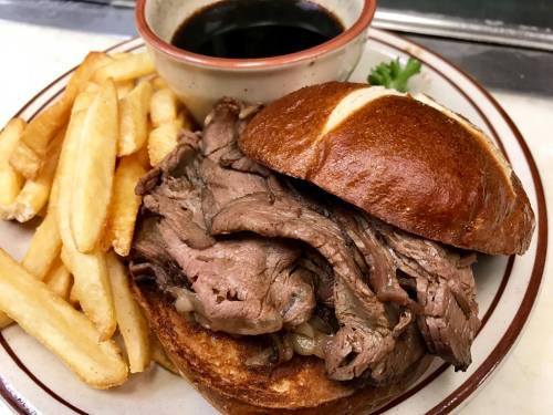 A hearty roast beef sandwich on a pretzel bun, served with fries and a small bowl of dipping sauce.