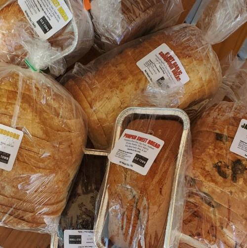 A variety of loaves of bread and sweet bread, all wrapped in plastic with labels, displayed on a wooden surface.