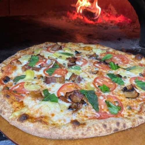 A freshly baked pizza topped with tomatoes, spinach, artichokes, and bacon, with a wood-fired oven in the background.