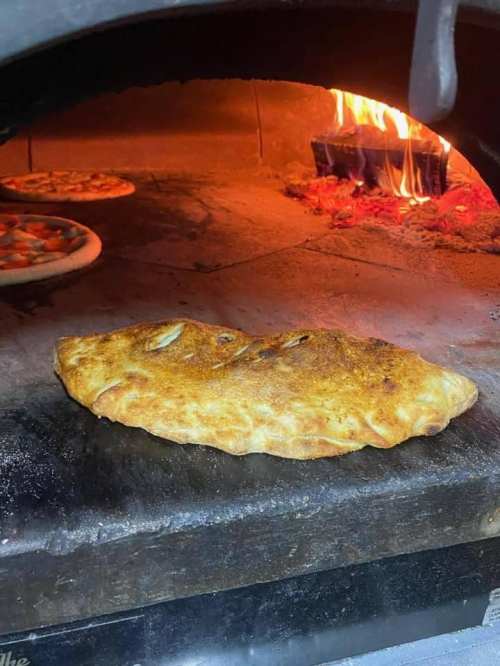 A freshly baked calzone resting on a stone surface in a wood-fired oven, with pizzas visible in the background.