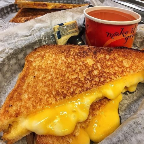 A golden-brown grilled cheese sandwich with melted cheese, served with a cup of soup and condiments on a tray.