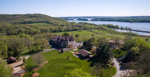 Aerial view of a large house surrounded by lush greenery and a river in the background.