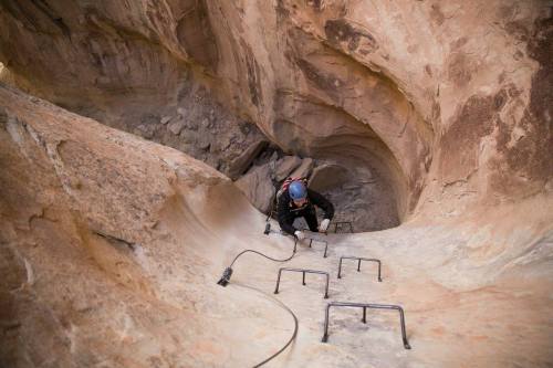 A climber ascends a steep rock face using metal rungs in a narrow canyon.