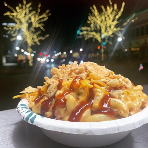 A close-up of a bowl of mac and cheese topped with barbecue sauce and crispy onions, with festive lights in the background.