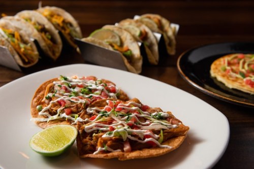 Two tacos topped with meat, vegetables, and sauce, served with a lime wedge, with more tacos in the background.