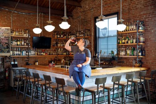 A bartender sits on a bar stool, enjoying a drink in a rustic bar with brick walls and shelves of liquor.