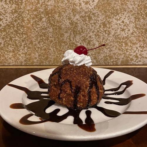 A dessert featuring a fried ice cream ball topped with whipped cream, a cherry, and drizzled with chocolate sauce.