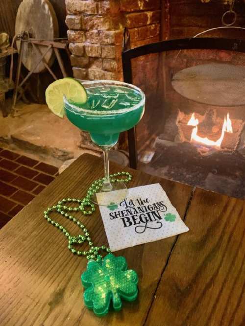 A green cocktail with a lime slice sits on a wooden table next to a St. Patrick's Day decoration and a fireplace.