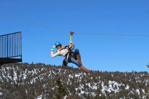 A person in a blue helmet zip-lining against a clear blue sky, smiling with a scenic mountain backdrop.