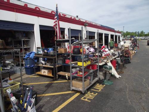 A cluttered outdoor market with various items for sale, including tools, equipment, and boxes, in front of storage units.