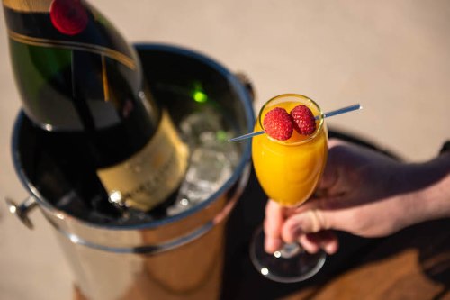 A hand holds a glass of orange drink garnished with raspberries, next to a bucket of champagne on ice.