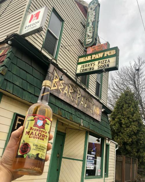 A hand holds a bottle of Jeppson's Malört in front of the Bear Paw Inn, with a sign announcing Jerry's Pizza coming soon.