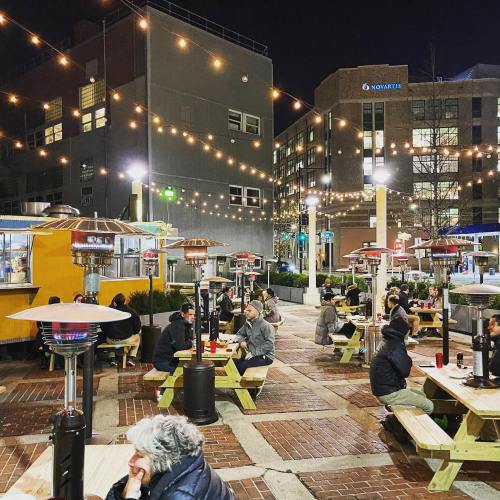 Outdoor dining area at night, featuring string lights, heaters, and people seated at picnic tables.