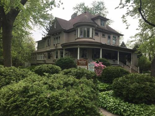 A large, historic house surrounded by lush greenery and blooming flowers, featuring a wraparound porch.