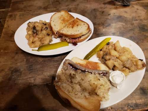 Two plates of food: one with a sandwich, pickles, and apple sauce; the other with sauerkraut and a toasted sandwich.