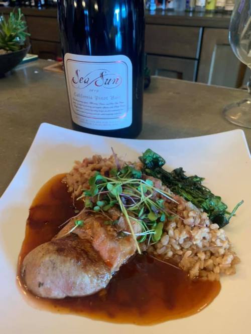 A plate featuring a slice of meat with sauce, grains, sautéed greens, and a bottle of Sea Sun California Pinot Noir.