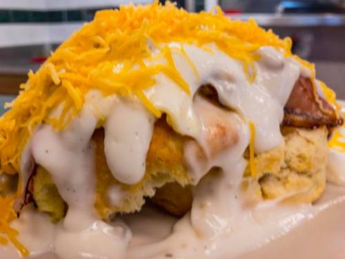A close-up of a biscuit topped with creamy gravy, ham, and a generous layer of shredded cheese.