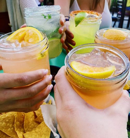 A group of friends toasting with colorful drinks in mason jars, surrounded by tortilla chips on a table.