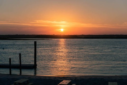 A serene sunset over a calm waterway, with silhouettes of posts and a sandy shore in the foreground.