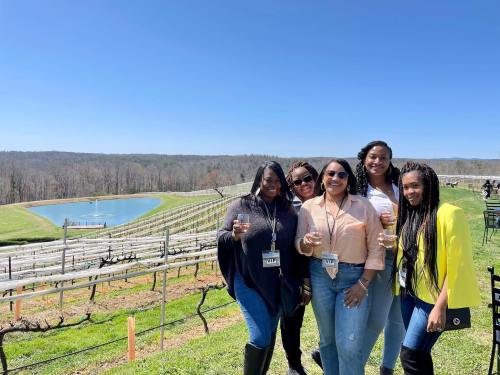 A group of five women holding drinks, smiling outdoors at a vineyard with a scenic view of a lake and hills.