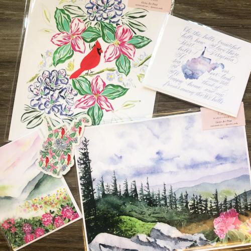 A collection of watercolor art prints featuring flowers, a cardinal, and a mountain landscape on a wooden surface.