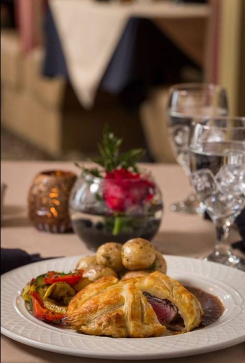A plated meal featuring beef Wellington, roasted vegetables, and potatoes, with elegant table settings in the background.