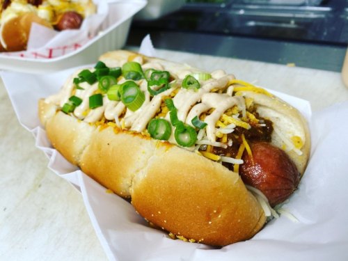 A close-up of a hot dog topped with cheese, green onions, and sauce, served in a paper tray.