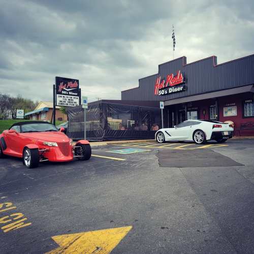 Two stylish cars parked outside a retro diner, with a cloudy sky overhead and a vibrant sign reading "Hot Rods 50's Diner."