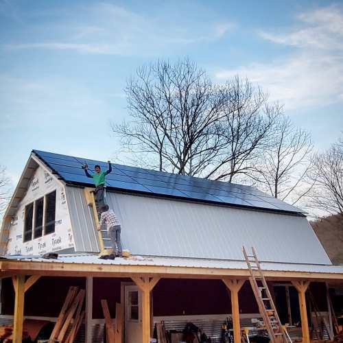 Two workers install solar panels on a barn roof, with trees and a blue sky in the background.