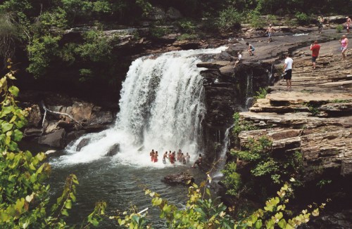 A waterfall cascades into a pool, with people swimming and enjoying the surrounding rocky landscape and greenery.