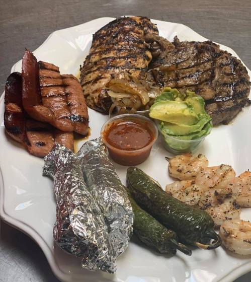 A plate featuring grilled chicken, steak, shrimp, sausage, jalapeños, avocado, and a side of sauce in a foil wrap.