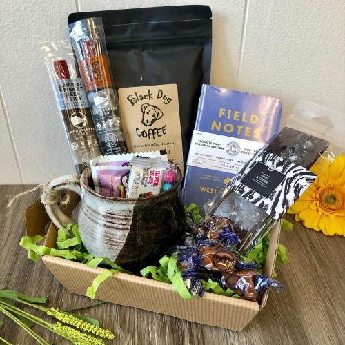 A gift basket featuring coffee, snacks, a notebook, and a handmade mug, arranged with decorative filler.